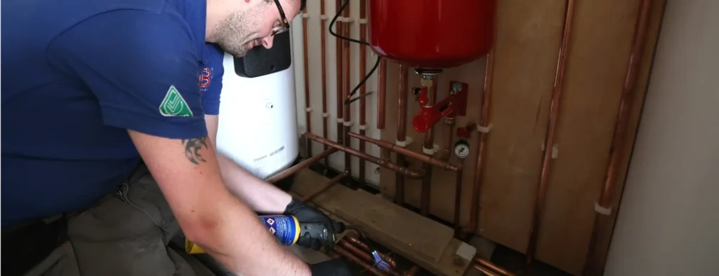 A person wearing a blue shirt and black gloves uses a handheld tool to work on copper pipes in the mechanical room, tending to boiler maintenance. Red and white machinery are visible in the background.