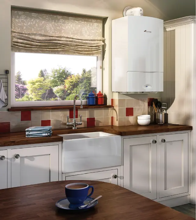 Modern kitchen with a wooden countertop, white cabinetry, a farmhouse sink, a Worcester wall-mounted boiler, and a window with a view of trees. A cup of coffee sits on the table in the foreground.