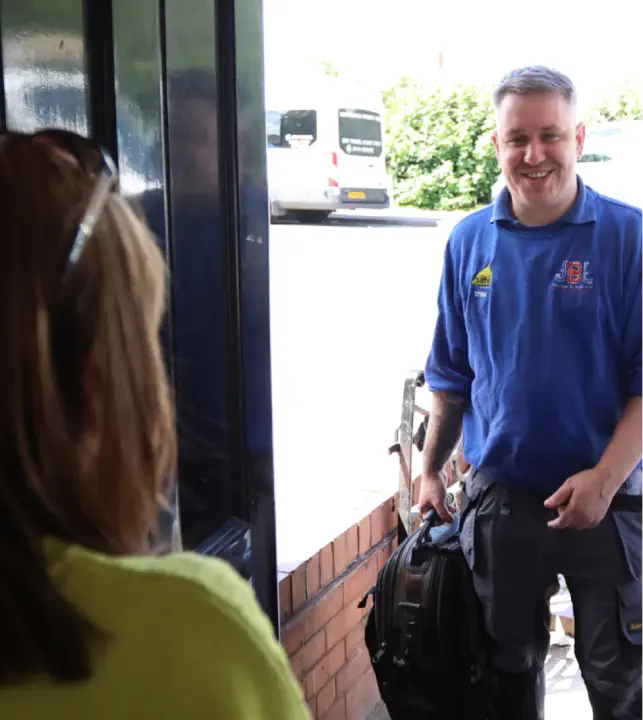 A person with grey hair in a blue shirt is smiling and carrying a black bag. They are at a doorstep, engaging with a person wearing glasses and a yellow shirt. A Worcester Boilers service van is parked nearby on this sunny day.